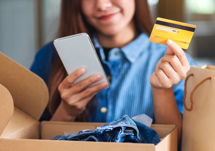 Una mujer joven está en el proceso de realizar una compra, sosteniendo una tarjeta de crédito amarilla y un smartphone blanco, con un cajón lleno de ropa y una bolsa de compras a su lado.