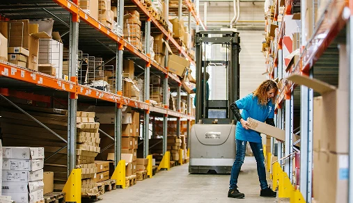Una trabajadora en un almacén, moviendo cajas de cartón, rodeada de otros productos y un forklift en el fondo.