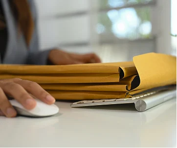 Persona sentada en una mesa, sosteniendo un ratón de computadora blanco y un archivo amarillo abierto, con un escritorio blanco y un archivo de archivo de metal en el fondo.