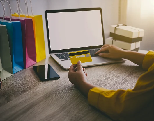 Persona sentada en una mesa, utilizando una laptop y una tarjeta de crédito, con bolsas de compras en el fondo.