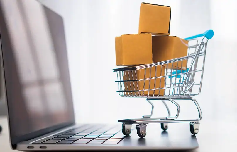 Una cesta de compras llena de cajas de cartón marrón sobre una computadora portátil con una pantalla abierta.