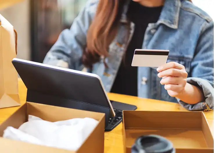 Una mujer joven sentada en una oficina, con una laptop y una tarjeta de crédito en la mano, rodeada de un entorno de trabajo casual.