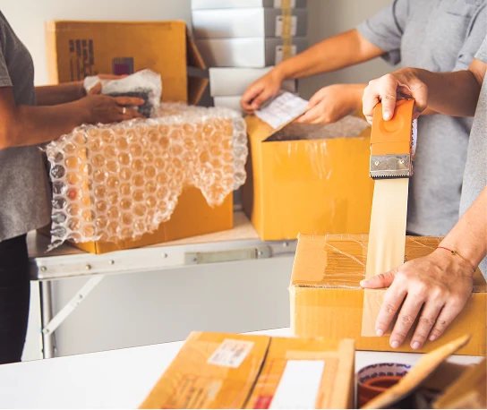 Tres personas empaquetando cajas en una mesa con fondo blanco.