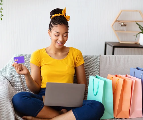 Una mujer joven sentada en un sofá, sosteniendo una laptop y una tarjeta de crédito, sonriendo y aparentemente disfrutando de su tiempo, rodeada de bolsas de compras.